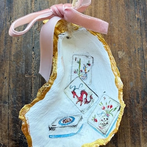 May include: Hand-painted oyster shell ornament with gold trim and a pink velvet ribbon. The shell features miniature illustrations of playing cards with floral and dragon designs. A unique, decorative item.
