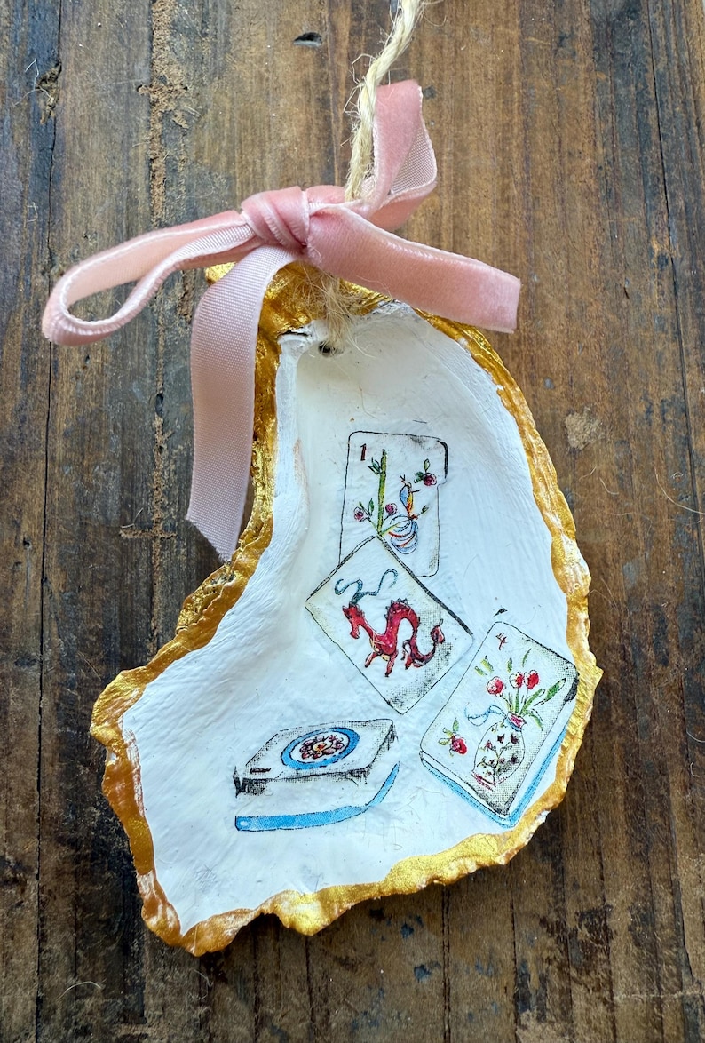 May include: Hand-painted oyster shell ornament with gold trim and a pink velvet ribbon. The shell features miniature illustrations of playing cards with floral and dragon designs. A unique, decorative item.