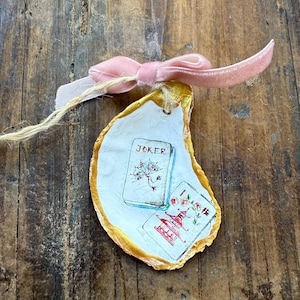 May include: Hand-painted oyster shell with gold trim, featuring playing card designs and the word "JOKER." A pink velvet bow and twine are attached. The shell rests on a rustic wooden surface.