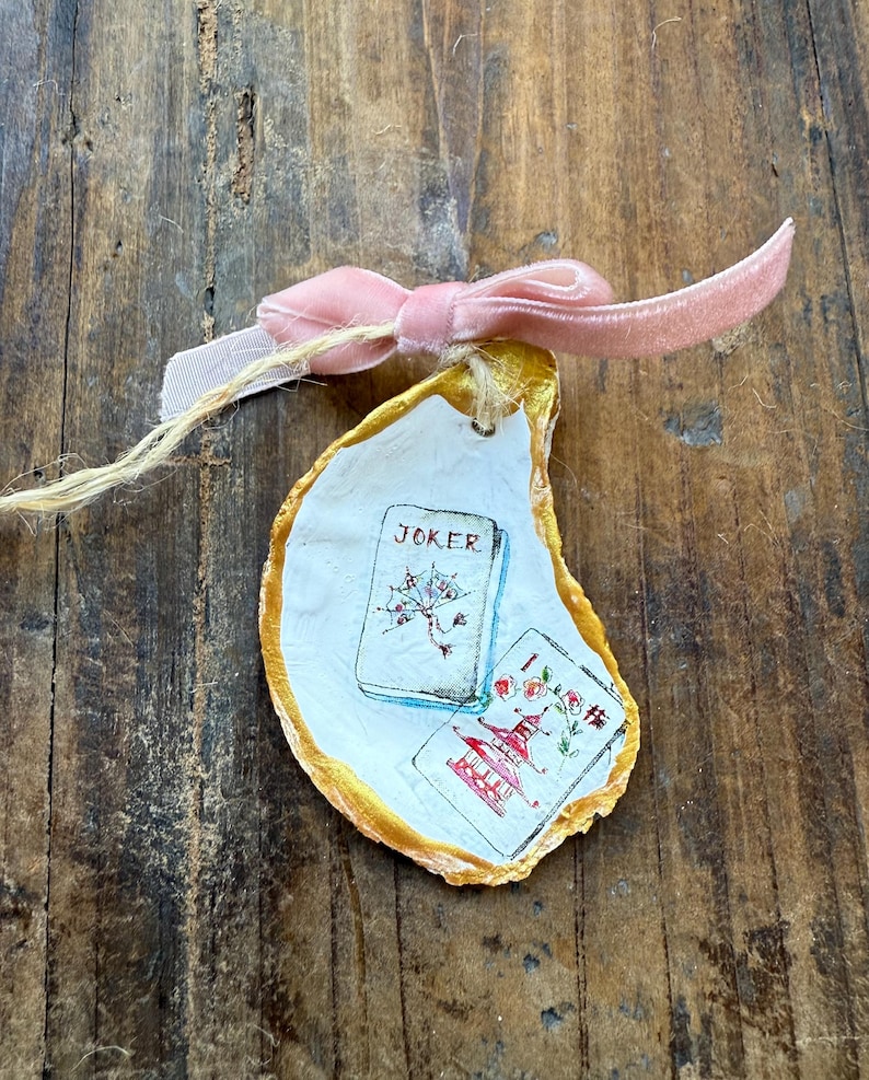 May include: Hand-painted oyster shell with gold trim, featuring playing card designs and the word "JOKER." A pink velvet bow and twine are attached. The shell rests on a rustic wooden surface.
