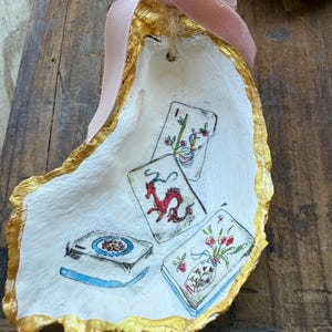 May include: Hand-painted oyster shell ornament with a gold gilded edge and a pink velvet ribbon. The shell features miniature illustrations of playing cards with a dragon, flowers, and other designs. A unique, decorative item.