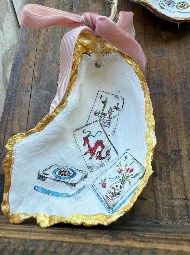 May include: Hand-painted oyster shell ornament with a gold gilded edge and a pink velvet ribbon. The shell features miniature illustrations of playing cards with a dragon, flowers, and other designs. A unique, decorative item.