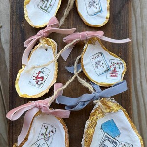 May include: Hand-painted oyster shells with gold edges, featuring playing card designs and tied with pink and blue velvet ribbons. The shells are displayed on a rustic wooden surface, creating a unique, handcrafted aesthetic.