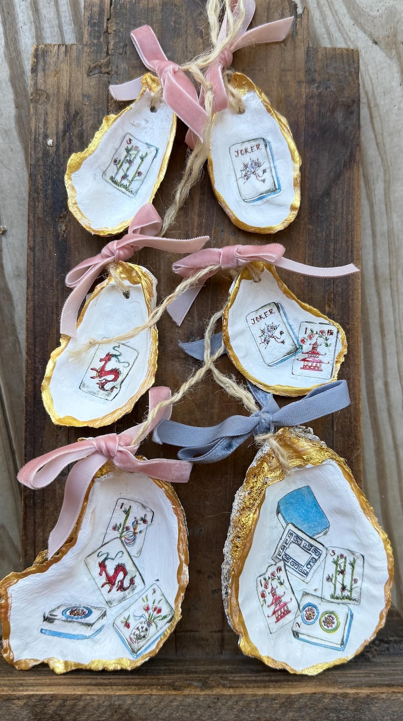 May include: Hand-painted oyster shells with gold edges, featuring playing card designs and tied with pink and blue velvet ribbons. The shells are displayed on a rustic wooden surface, creating a unique, handcrafted aesthetic.