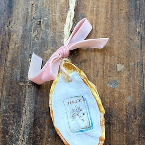 May include: An oyster shell ornament with a gold-painted edge, a white interior, and a small card with the word "JOKER" and a floral illustration. A pink velvet ribbon and natural twine are attached for hanging.