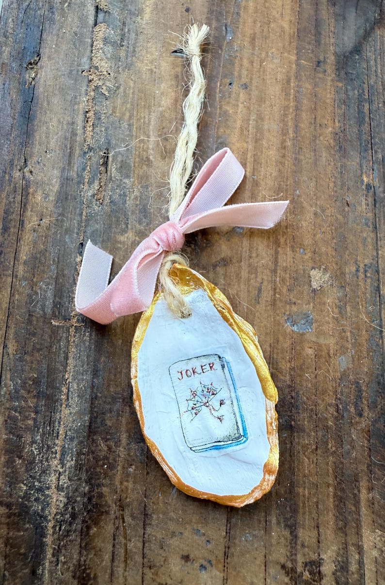 May include: An oyster shell ornament with a gold-painted edge, a white interior, and a small card with the word "JOKER" and a floral illustration. A pink velvet ribbon and natural twine are attached for hanging.