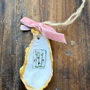 May include: An oyster shell ornament with a gold-painted edge, a white interior, and a small floral illustration. A pink velvet ribbon is tied in a bow at the top, and a natural twine loop is attached for hanging.