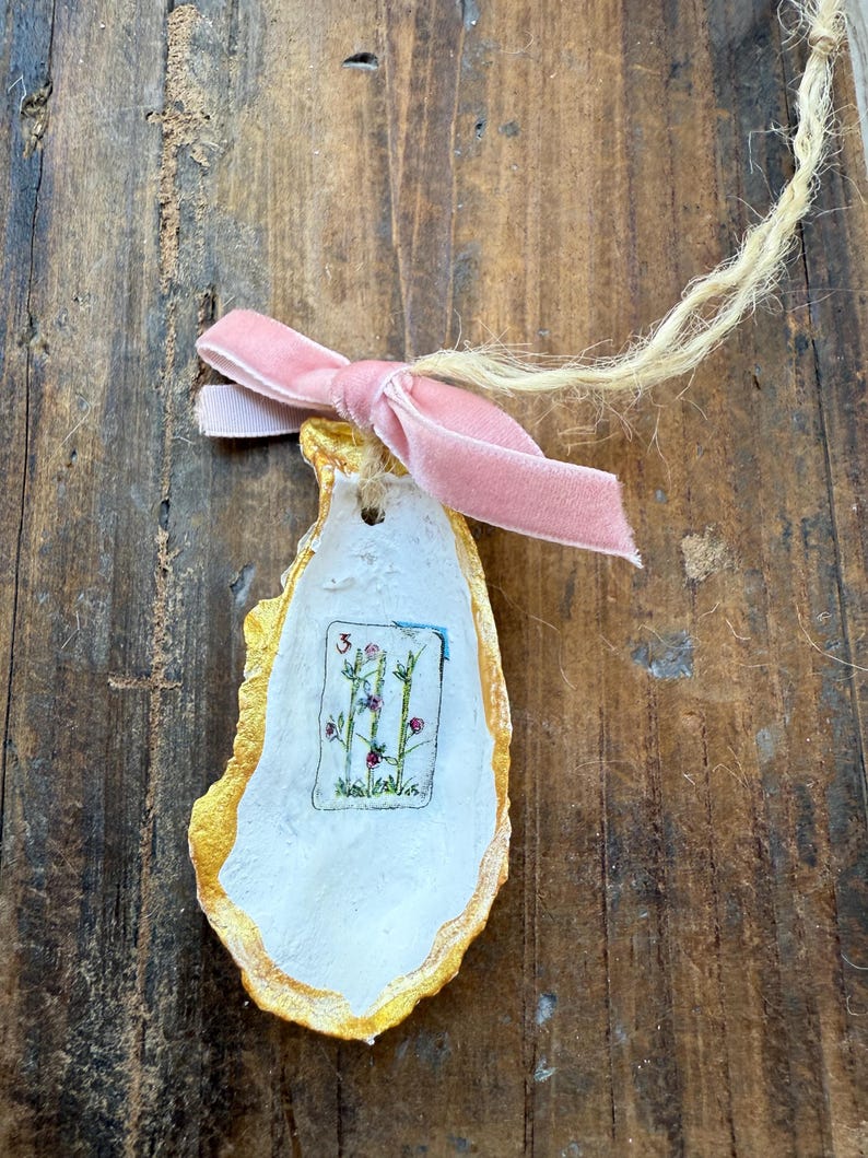 May include: An oyster shell ornament with a gold-painted edge, a white interior, and a small floral illustration. A pink velvet ribbon is tied in a bow at the top, and a natural twine loop is attached for hanging.