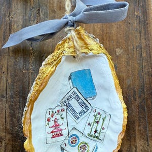May include: Hand-painted oyster shell ornament with gold trim and a light blue ribbon. The shell features a Mahjong tile design with red, blue, and green accents. A unique, handcrafted decorative item.