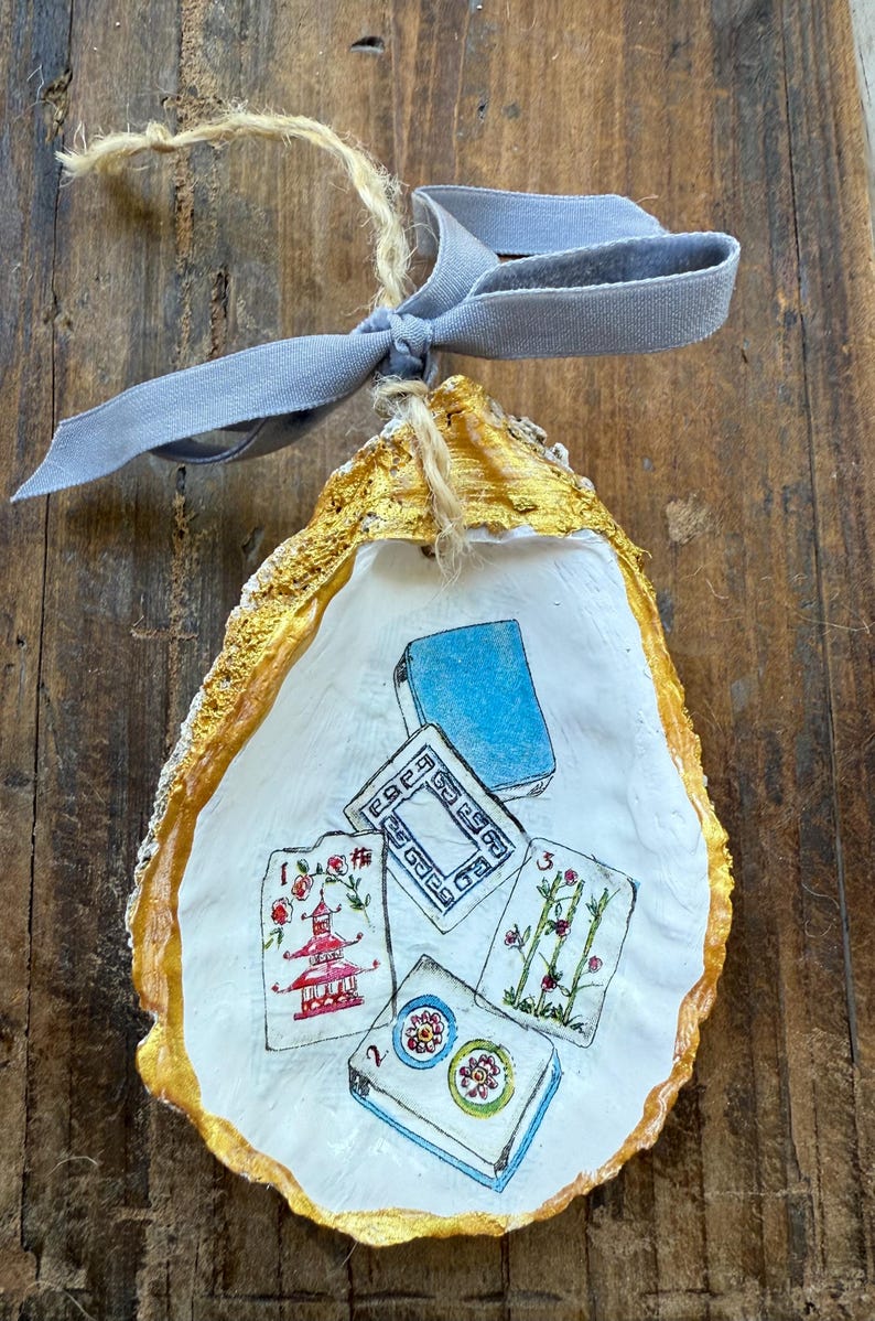 May include: Hand-painted oyster shell ornament with gold trim and a light blue ribbon. The shell features a Mahjong tile design with red, blue, and green accents. A unique, handcrafted decorative item.