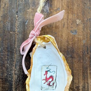 May include: An oyster shell ornament with a gold-painted edge and a pink velvet ribbon. The shell features a small, framed illustration of a red dragon. A natural twine loop is attached for hanging.