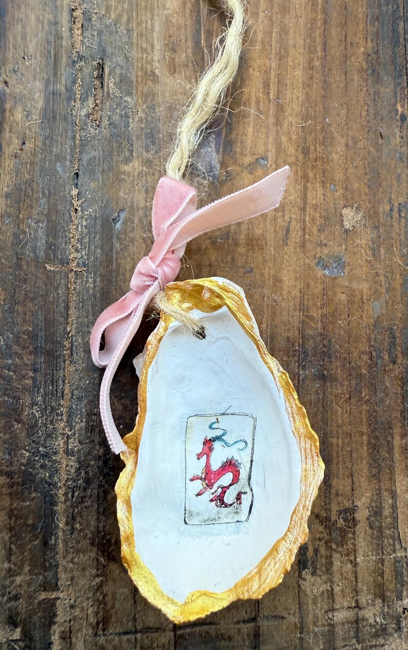 May include: An oyster shell ornament with a gold-painted edge and a pink velvet ribbon. The shell features a small, framed illustration of a red dragon. A natural twine loop is attached for hanging.