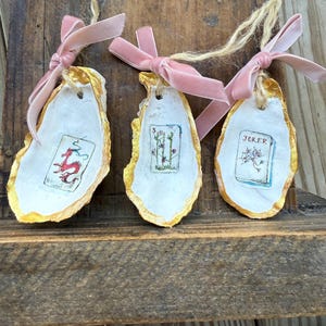 May include: Three oyster shell ornaments with gold edges, white interiors, and pink velvet ribbon bows. Each shell features a small, hand-painted playing card design: a red dragon, a floral arrangement, and a "Joker" card.