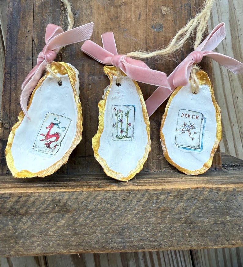 May include: Three oyster shell ornaments with gold edges, white interiors, and pink velvet ribbon bows. Each shell features a small, hand-painted playing card design: a red dragon, a floral arrangement, and a "Joker" card.