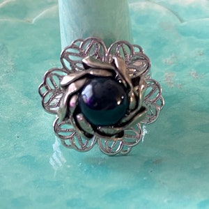 May include: A silver-toned ring with a dark blue, possibly black, center stone. The ring features a floral design with openwork petals and leaves surrounding the central stone. The ring is displayed on a light blue surface.