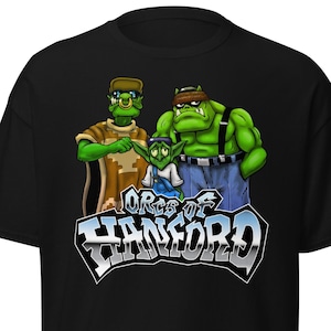 May include: A black short-sleeve t-shirt featuring a graphic of three green orc-like characters. Below the characters, the text reads "ORCS OF HANFORD" in a bold, metallic-style font with a blue gradient and a thick black outline.
