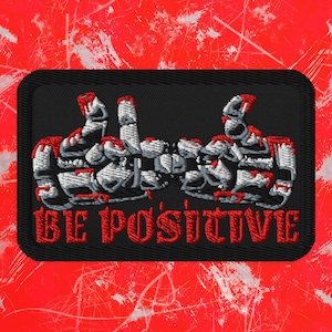 May include: Embroidered patch with a black background and a red border. The patch features two thumbs up in white and gray, with red accents. The words "BE POSITIVE" are embroidered in red.