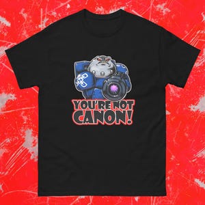 May include: Black t-shirt featuring a cartoon illustration of a blue and gray armored figure holding a purple lens. The text "YOU'RE NOT CANON!" is displayed in red and white. The shirt is made of a soft, comfortable material.