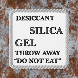 May include: A white rectangular patch with black embroidered text that reads "DESICCANT SILICA GEL THROW AWAY 'DO NOT EAT'". The patch has a gray stitched border and is set against a rusty brown background.