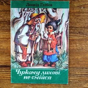May include: A vintage children's book cover featuring a colorful illustration of an older man with a white beard and a young boy holding a bird in a forest setting. The book title is "Chukomu lihovi ne smiisa" in Cyrillic script.