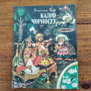May include: A vintage children's book cover with a dark green background. The title is "Kalif-Chornoguz" in Cyrillic script. The illustration depicts a scene with three figures, a peacock, and a bird in a cage.