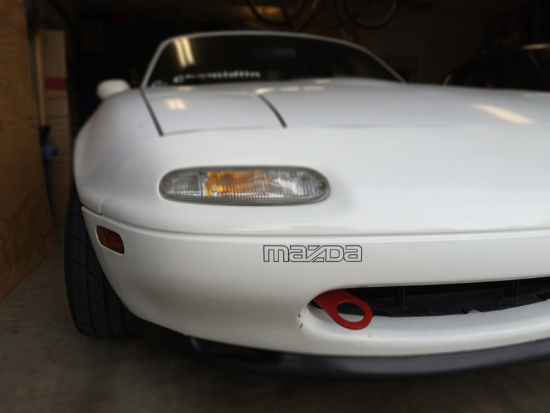May include: White Mazda Miata sports car with a red tow hook. The car has a black bumper and a black grill. The Mazda logo is visible on the front of the car.