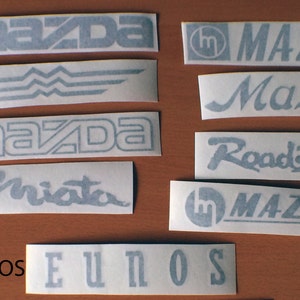 May include: A collection of eight different black and white vinyl decals featuring the Mazda logo and various text, including "SOLID", "OUTLINE", "MIATA", "EUNOS", "Roadster", "Mazda", and "1959".