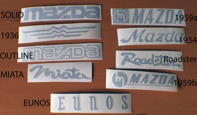 May include: A collection of eight different black and white vinyl decals featuring the Mazda logo and various text, including "SOLID", "OUTLINE", "MIATA", "EUNOS", "Roadster", "Mazda", and "1959".