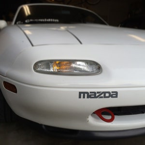 May include: A white Mazda Miata sports car with a red tow hook. The car has a black bumper and a black grill. The car has a yellow turn signal on the front.