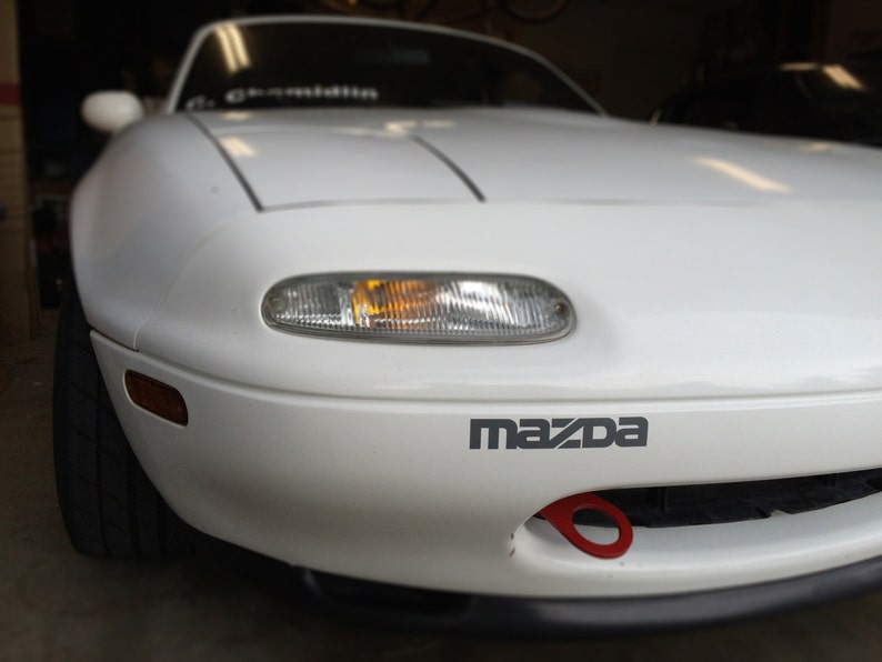 May include: A white Mazda Miata sports car with a red tow hook. The car has a black bumper and a black grill. The car has a yellow turn signal on the front.