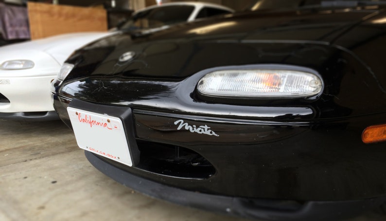 May include: A black Miata sports car with a California license plate. The car has a silver Miata emblem on the front bumper.