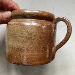 May include: A brown ceramic mug with a rounded handle. The mug has a textured surface and a slightly flared rim. The color is a warm, earthy brown with variations in tone. The mug appears to be handmade.