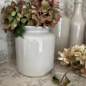 May include: A white ceramic jar with the embossed text LAB printed on the side, containing a cluster of dried pink and green hydrangea flowers. The jar has a cylindrical shape with a short neck and a smooth, glossy finish, standing upright.