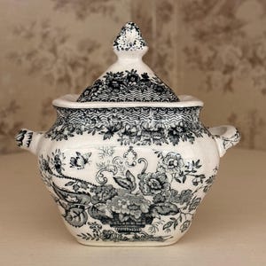 May include: A cream-coloured ceramic sugar bowl with a matching lid, featuring a detailed black floral transferware pattern. The vessel has two side handles and a pointed finial on the lid, decorated with intricate botanical and scroll motifs.