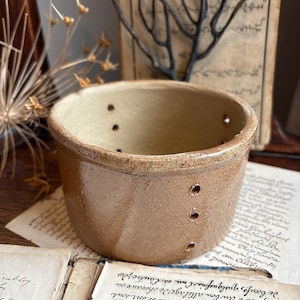 May include: A small, round, tan ceramic bowl with several small holes around the side. The bowl has a slightly glossy finish and sits on old, handwritten papers. The background includes dried plants and a vintage book.