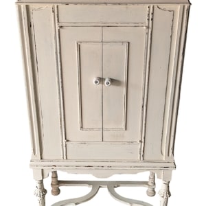 May include: Off-white, distressed cabinet with two doors and white knobs. The cabinet has a rectangular shape with decorative molding and stands on four ornate legs. The cabinet is painted with a distressed finish, giving it a vintage look.