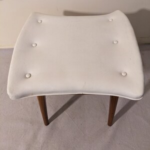 May include: White upholstered stool with a curved top and four button tufts. The stool has wooden legs.