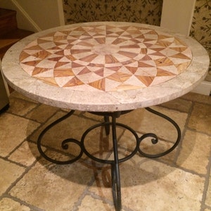 May include: A round, stone table top with a geometric pattern of light and dark brown tiles. The table has a black wrought iron base with a scrolled design.