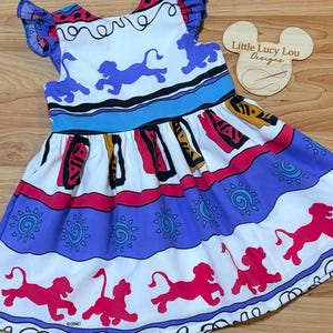 May include: A white dress with a colorful print featuring purple lions, suns, and geometric patterns. The dress has a blue neckline and ruffled sleeves. The Disney logo is visible on the bottom hem. The dress is designed for children.