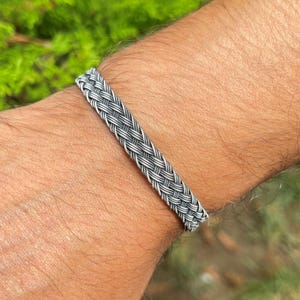 May include: A silver-coloured braided bracelet. The bracelet is made of interwoven metal strands, creating a textured, woven appearance. The bracelet is worn on a wrist, showcasing the intricate design and craftsmanship.