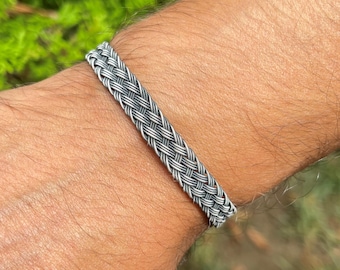 Op de afbeelding: Een zilverkleurige gevlochten armband. De armband is gemaakt van verweven metalen strengen, waardoor een getextureerde, geweven uitstraling ontstaat. De armband wordt om een pols gedragen en toont het ingewikkelde ontwerp en vakmanschap.