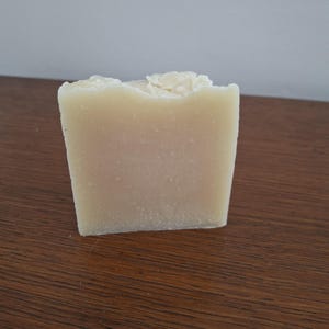 Può includere: Una saponetta rettangolare fatta a mano. Il sapone è di colore beige chiaro con una superficie leggermente strutturata. Il bordo superiore ha un aspetto ruvido e irregolare. Il sapone è su una superficie di legno marrone scuro.