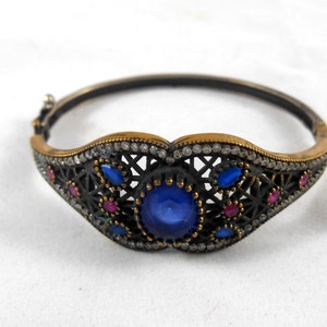 May include: A silver bangle bracelet with a filigree design and blue and red gemstones. The bracelet is decorated with small, clear stones.