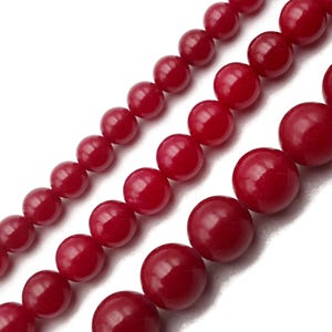 May include: Close-up of several strands of round, glossy, deep red beads. The beads vary slightly in size and are strung together, creating a textured and visually appealing pattern. The beads are against a white background.