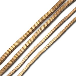 May include: Four strands of rectangular, metallic beads in a warm, copper color. The beads are uniform in size and shape, creating a textured, linear pattern. The strands are arranged diagonally against a white background.