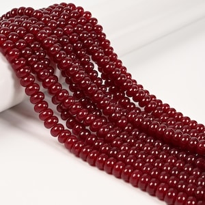 May include: A collection of multiple strands of deep red, smooth, round beads arranged in parallel lines. Each bead features a glossy finish and a uniform shape, creating a consistent texture across the entire set of strands displayed together.