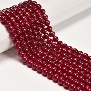 May include: Strands of glossy, deep red beads. The beads are perfectly round and uniform in size, creating a smooth, reflective surface. The beads are arranged in parallel rows, suggesting they are ready for jewelry making or crafting projects.
