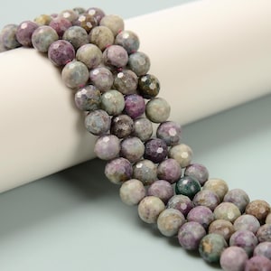 May include: Strands of faceted gemstone beads in shades of purple, gray, and green. The beads are round and have a textured surface. The beads are strung together, creating a necklace or bracelet.