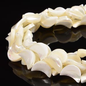 May include: A close-up of a necklace made of iridescent, off-white, crescent-shaped beads. The beads are strung together, creating a layered effect. The necklace is set against a black background, highlighting the pearlescent sheen of the beads.