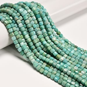 May include: Close-up of strands of turquoise-colored, faceted gemstone beads. The beads are cube-shaped and have a slightly irregular surface. The color varies from light to medium turquoise, with some beads showing hints of green. The beads are strung together, creating a flowing, textured appearance.